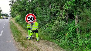 Deux hommes installant un panneau 90km/h sur une route départementale